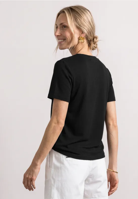 Frau von hinten in schwarzem T-Shirt und weißer Hose, blondes Haar im Dutt.