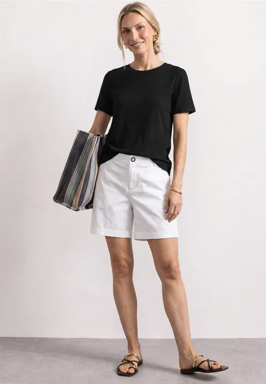 Frau trägt schwarzes T-Shirt, weiße Shorts, Sandalen und gestreifte Tasche.