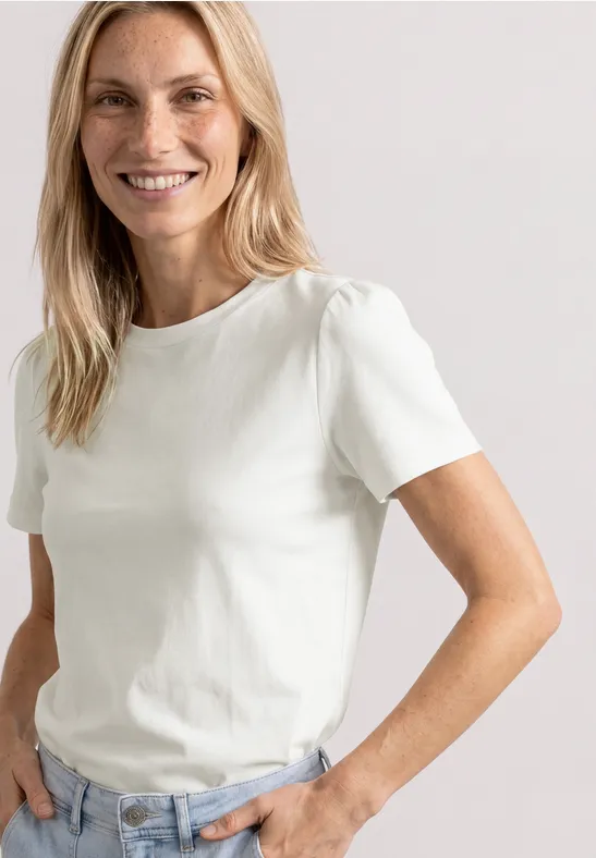 Frau in weißem T-Shirt und Jeans steht lässig mit einer Hand in der Tasche.