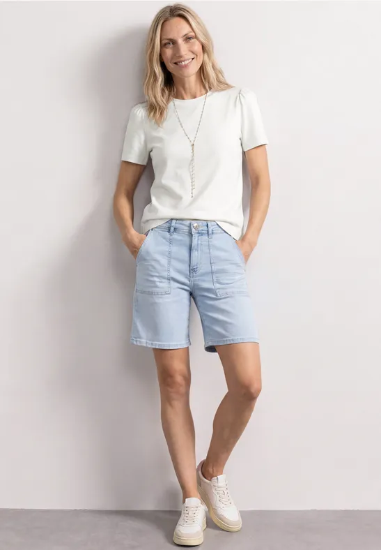Person in weißem T-Shirt, hellblauen Jeans-Shorts und weißen Sneakern steht entspannt vor weißer Wand.