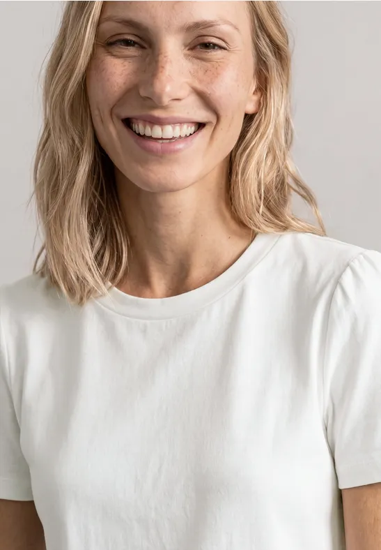 Eine Person mit schulterlangem, blondem Haar trägt ein schlichtes weißes T-Shirt.