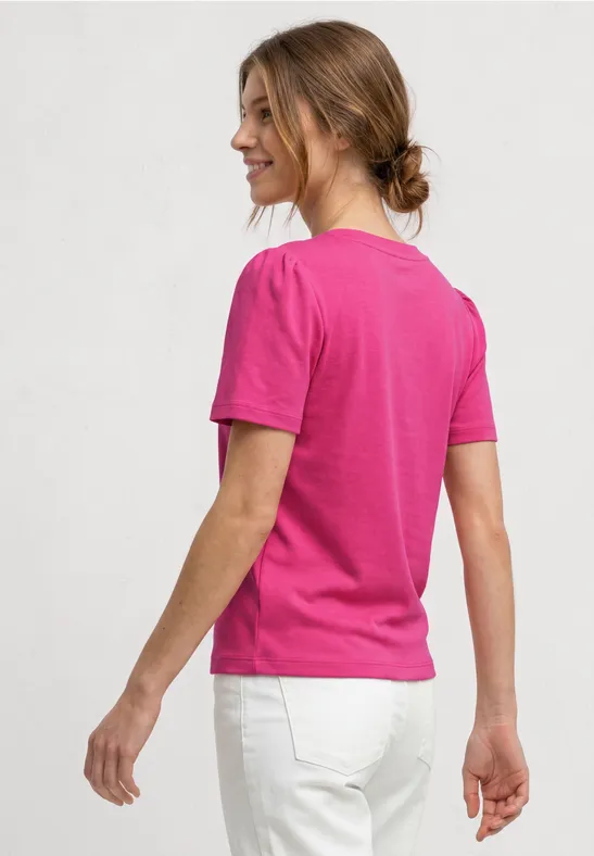 Een vrouw van achteren in een roze shirt en witte broek tegen een neutrale achtergrond.