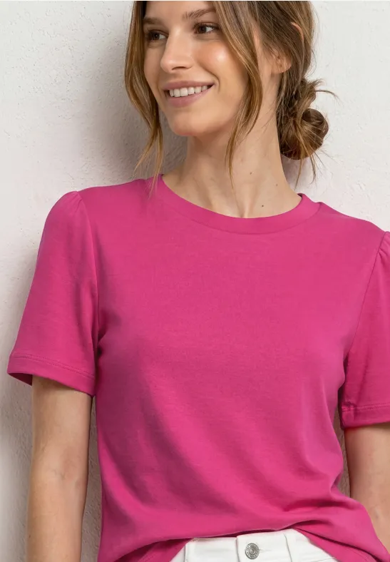 Vrouw in roze t-shirt en witte broek voor een lichte achtergrond, glimlachend.