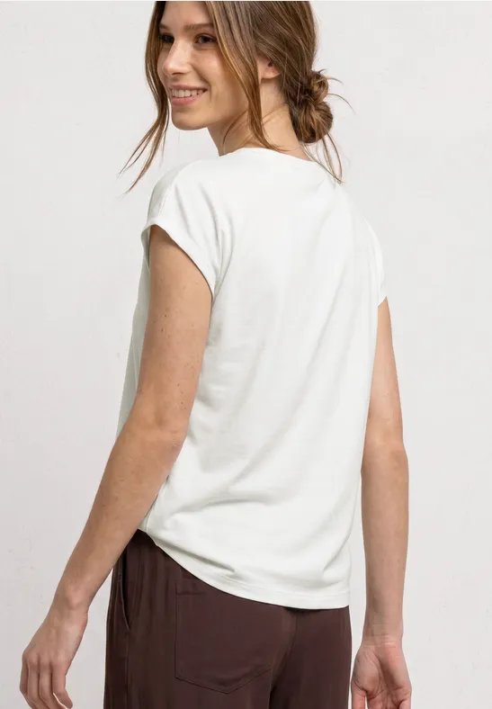 Vrouw van achteren in wit t-shirt en bruine broek tegen lichte achtergrond.