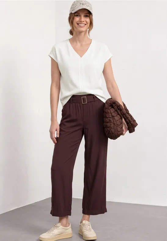 Vrouw in witte V-hals top, wijde bruine broek met riem, beige sneakers en gevlochten tas.