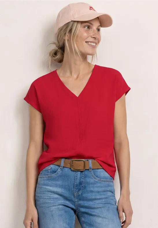 Frau in roter V-Bluse, Jeans und rosa Basecap, vor weißer Wand