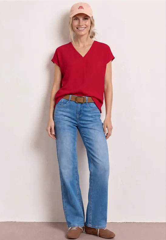 Frau in roter Bluse, blauer Jeans, braunem Gürtel und Stiefeletten vor hellem Hintergrund.