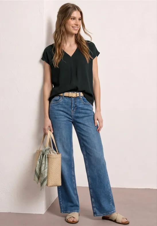 Vrouw in zwart V-hals T-shirt, blauwe rechte jeans, sandalen en strooien tas met sjaal, neutrale achtergrond.