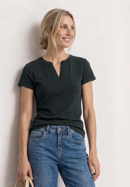 Vrouw in zwart t-shirt en blauwe spijkerbroek leunt ontspannen tegen een witte muur.