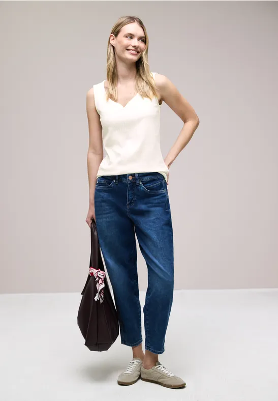 Frau in weißem Top, blauen Jeans und beigen Schuhen hält eine dunkelviolette Tasche mit Tuch, vor hellem Hintergrund.