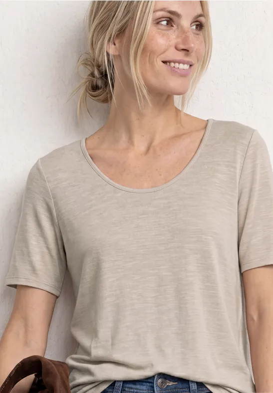 Eine Frau mit hochgestecktem Haar trägt ein schlichtes beiges Kurzarmshirt vor heller Wand.