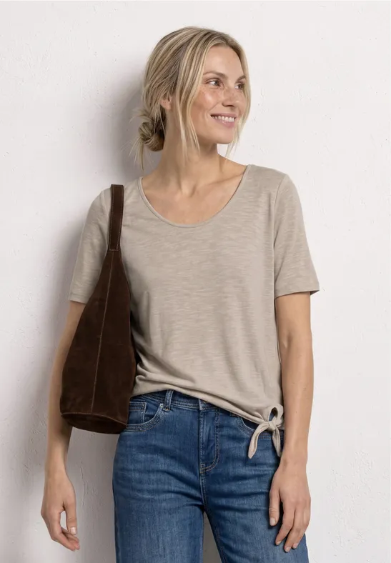 Frau trägt beiges, kurzärmeliges Shirt mit seitlicher Knoten-Optik, blaue High-Waist-Jeans und Schultertasche vor weißer Wand.