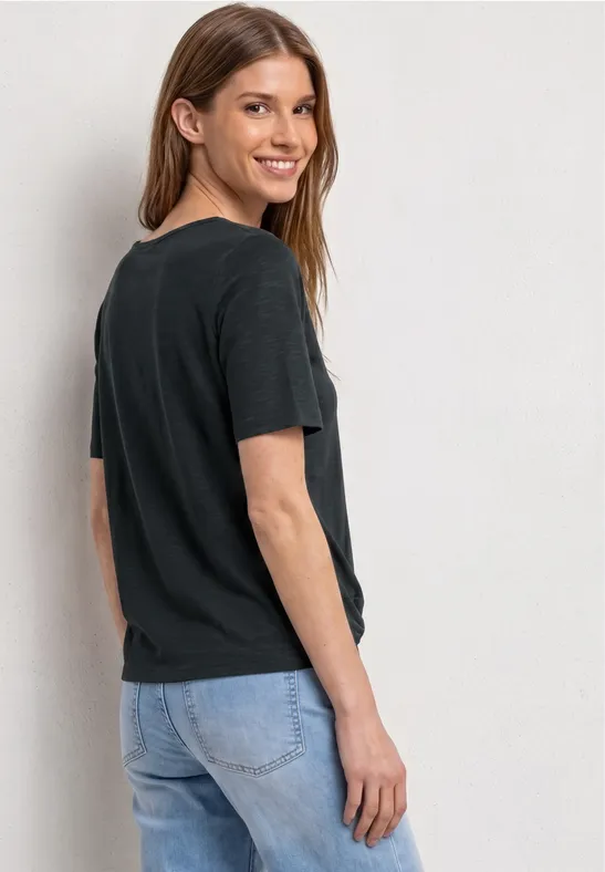 Frau im schwarzen T-Shirt und Jeans steht seitlich und blickt über die Schulter.