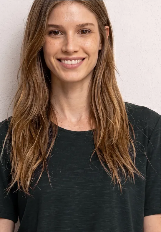 Eine Person mit langen braunen Haaren und schwarzem T-Shirt steht vor einer neutralen Wand.