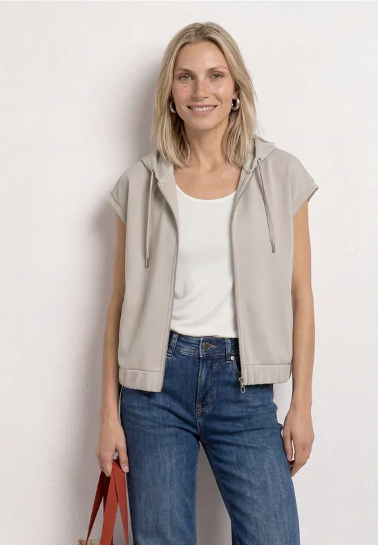 Frau mit ärmelloser Kapuzenweste, weißem Shirt, blauen Jeans und Handtasche.