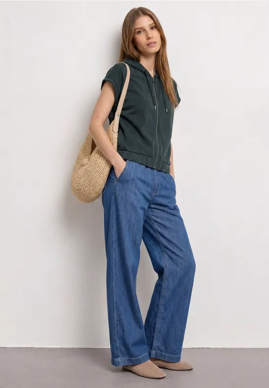 Vrouw in zwart capuchonshirt, wijde blauwe jeans, bruine schoenen en een strozak.
