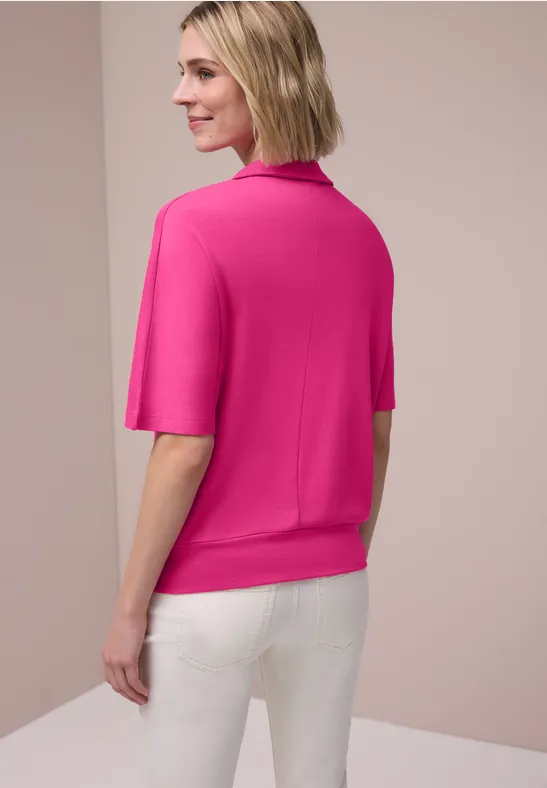 Een vrouw gezien van achteren voor een lichte achtergrond, met een roze kortemouwtrui en witte broek.