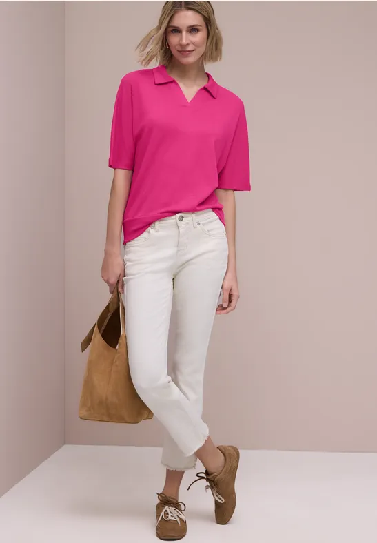 Een persoon voor een neutrale achtergrond draagt een roze poloshirt, witte jeans, bruine schoenen en houdt een tas vast.