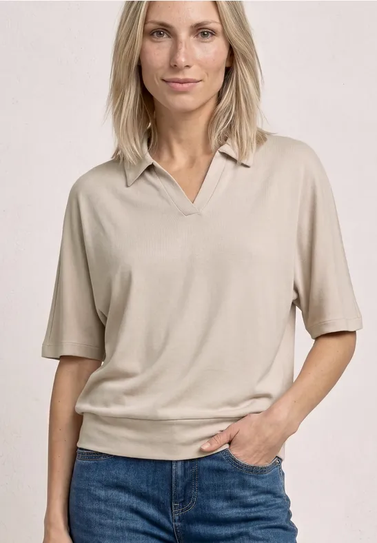 Vrouw draagt beige poloshirt met halflange mouwen en blauwe jeans, hand in de zak.