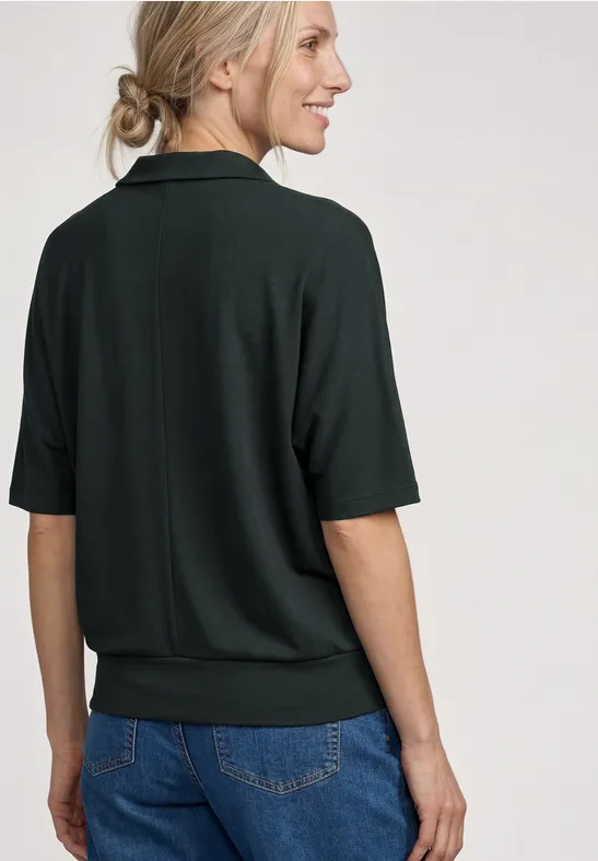 Vrouw van achteren in een donkergroen shirt met halflange mouwen en spijkerbroek, haar in een lage knot.