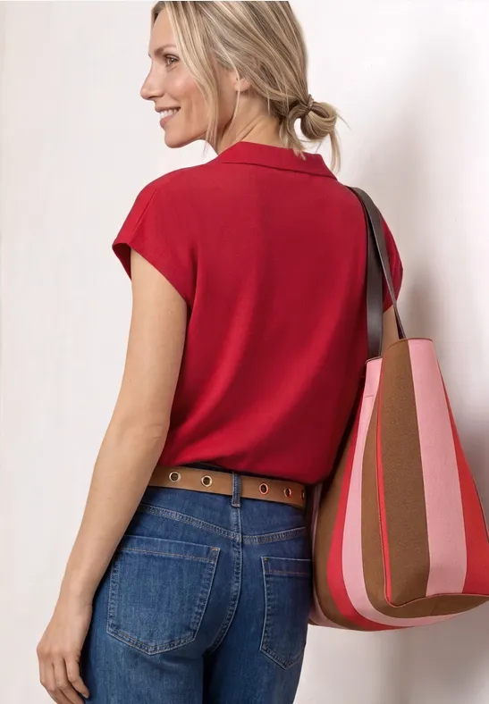 Een vrouw van achteren in een rood bloesje, spijkerbroek en riem draagt een gestreepte schoudertas.