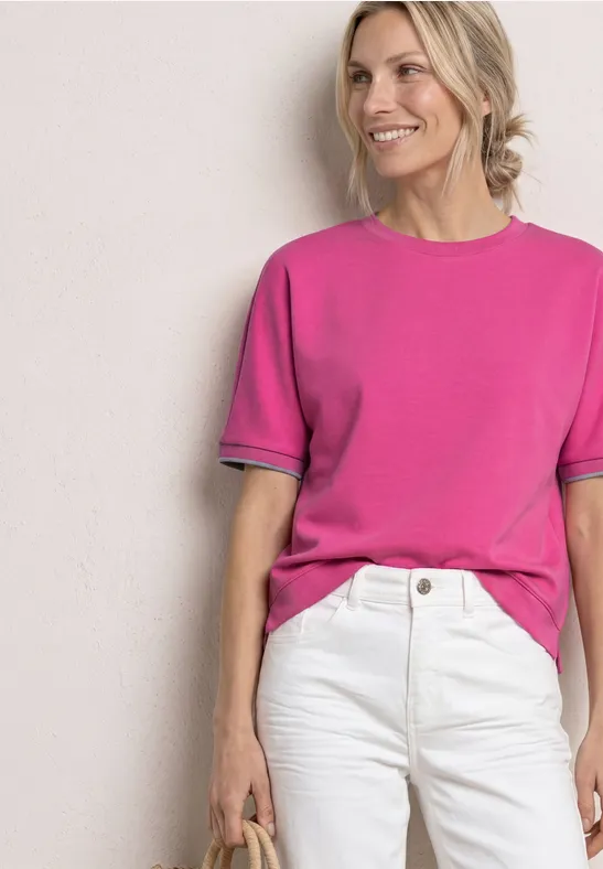 Frau in knallpinker T-Shirt und weißen Jeans, hält eine kleine Tasche vor neutralem Hintergrund.