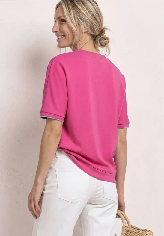 Eine Frau von hinten in pinkem Kurzarmshirt mit schwarzem Saum, weißen Hosen und geflochtener Korbtasche.