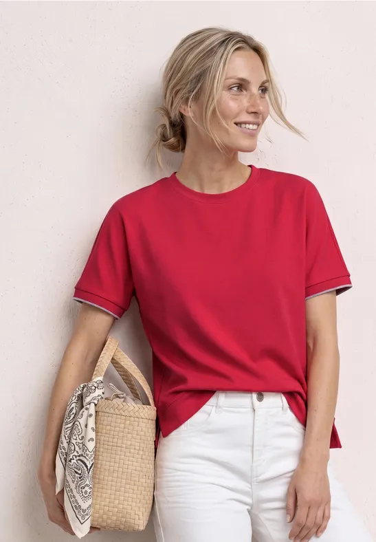Person trägt rotes T-Shirt, weiße Hose und hält Strohtasche vor neutralem Hintergrund.