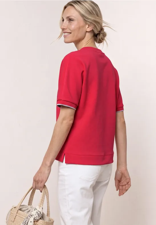 Frau von hinten in rotem Kurzarmshirt, weißer Hose und Korbtasche in der Hand.