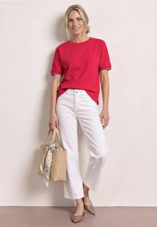 Frau in rotem T-Shirt, weißer Culotte, grauen Pumps, hält eine geflochtene Korbtasche mit Tuch.