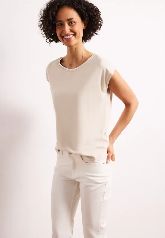 Vrouw in wit t-shirt en witte broek poseert voor een lichte achtergrond.