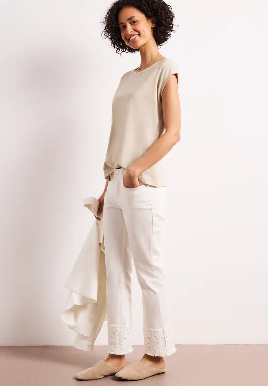 Vrouw met beige top en witte broek, houdt licht jasje vast tegen een witte achtergrond.