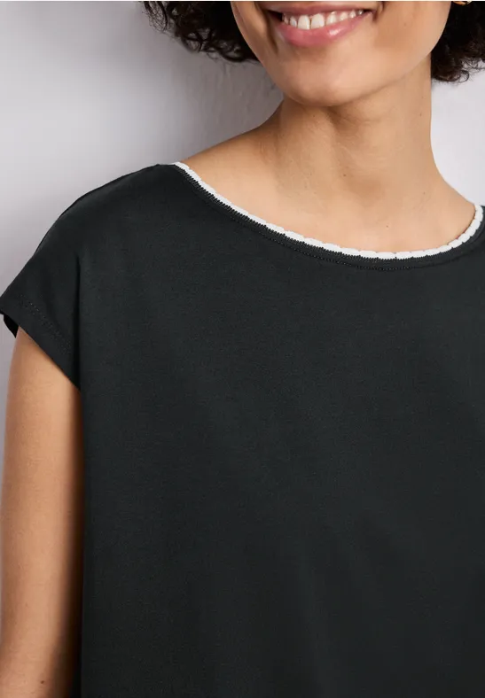 Eine Frau lächelt halb im Bild, trägt ein schlichtes schwarzes Oberteil mit weißem Rundhalsausschnitt.