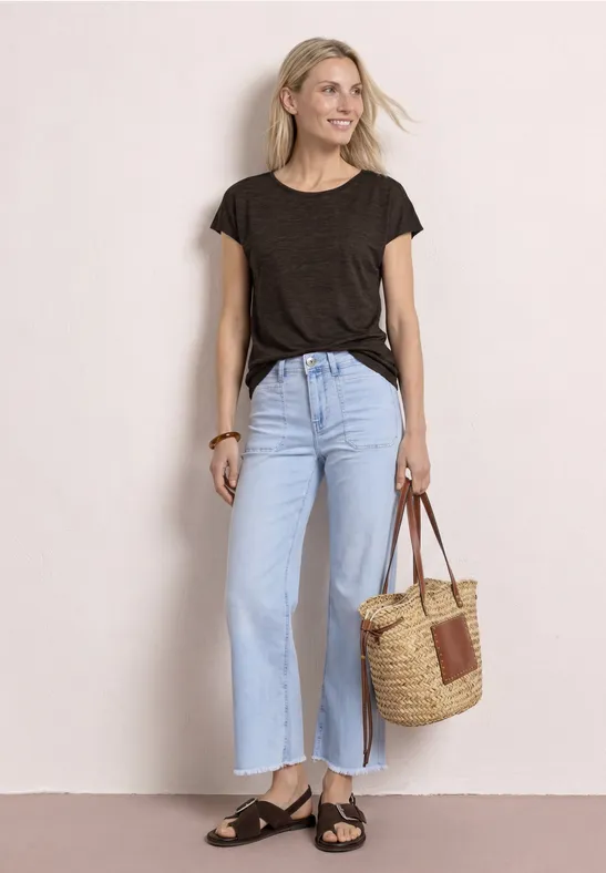 Frau in schwarzem T-Shirt, hellblauen, weitem Jeans, Sandalen und Basttasche vor weißem Hintergrund.