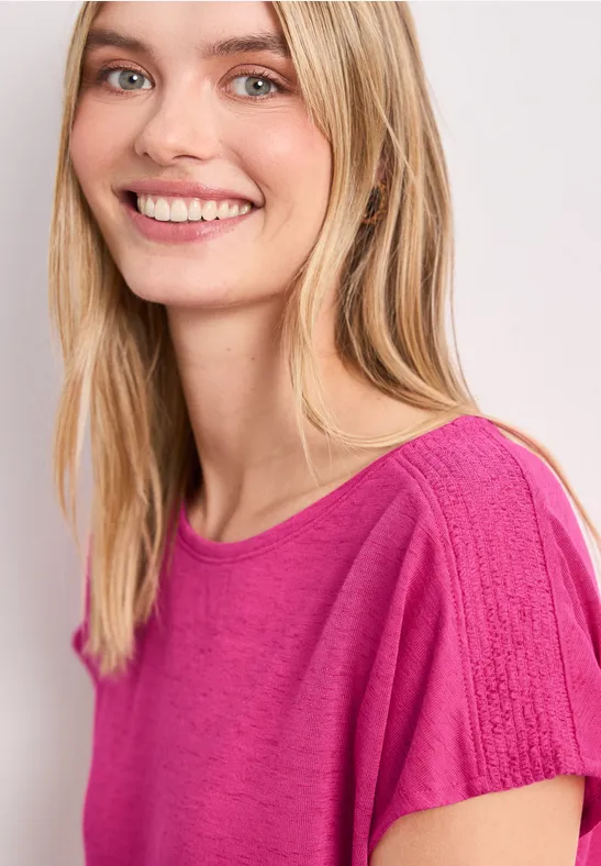 Person mit blondem Haar trägt pinkes Shirt vor hellem Hintergrund.
