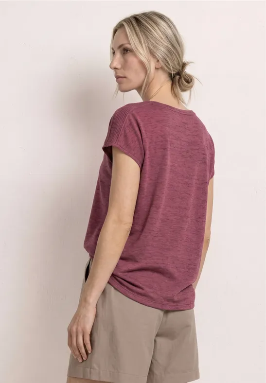 Frau steht in weinrotem T-Shirt und beigen Shorts, Rückenansicht, Blick abgewandt.
