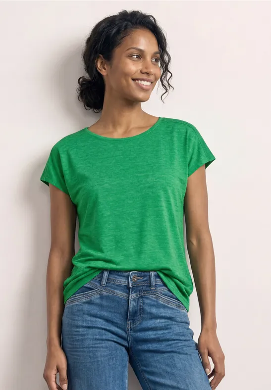 Femme portant un t-shirt vert et un jean bleu devant un fond blanc.