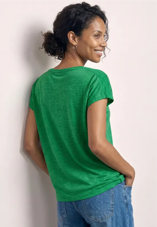 Une personne dos à la caméra, porte un t-shirt vert et un jean, les mains dans les poches.