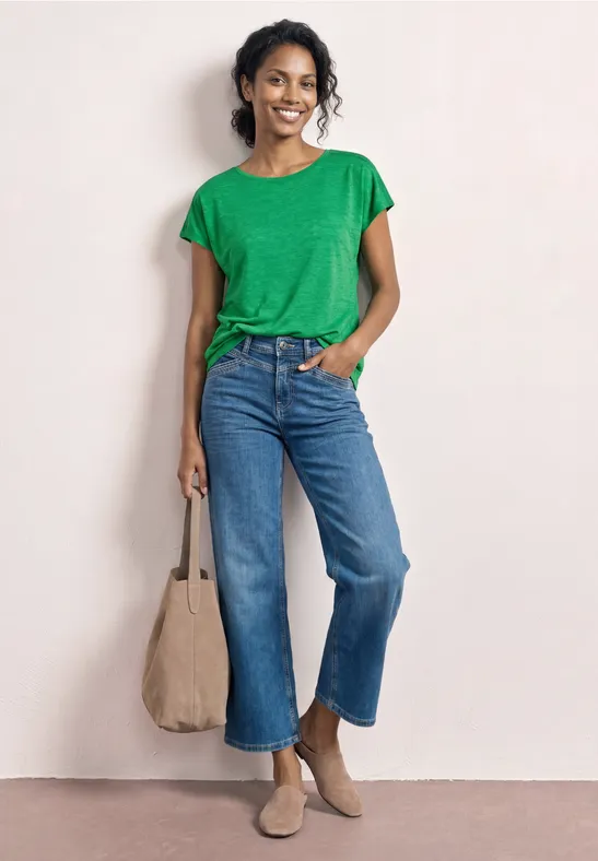 Femme portant un t-shirt vert, jean large, bottines et sac beiges, posant décontractée avec une main dans la poche.