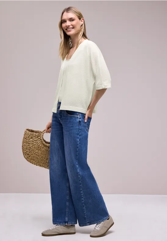 Frau in weiter blauer Jeans und weißer Bluse, Sneaker und geflochtene Strohtasche.