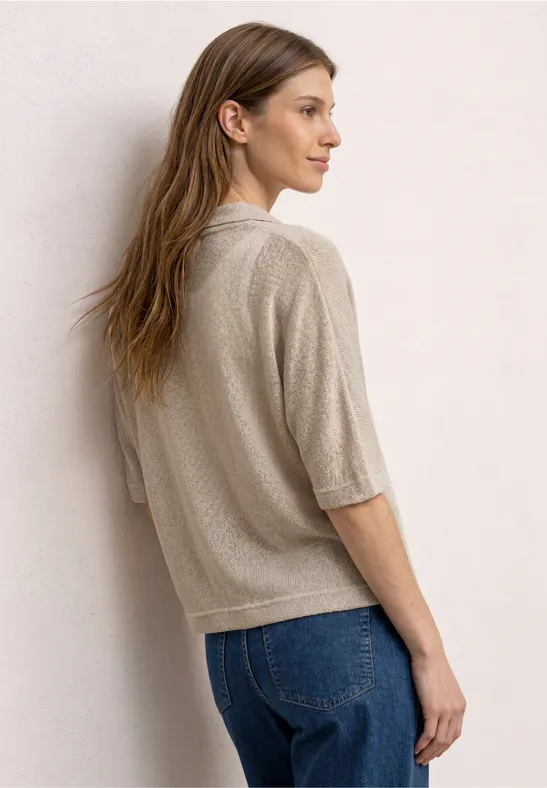 Frau steht mit dem Rücken zur Kamera, trägt beige Strickoberteil und blaue Jeans, vor heller Wand.