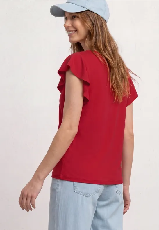 Frau steht vor hellem Hintergrund, trägt rote, kurzärmlige Bluse mit Volants, blaue Jeans und weiße Kappe.