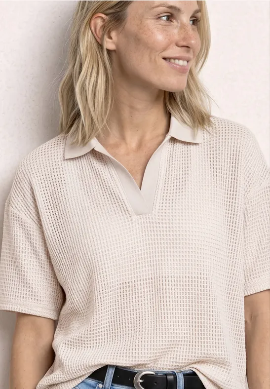 Vrouw in beige wafelpolo met korte mouwen, V-hals en zwarte riem, staand voor een lichte muur.