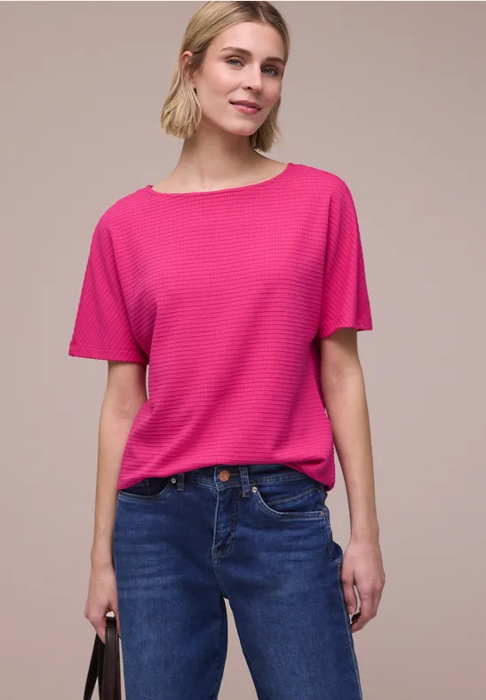 Persoon in roze T-shirt en blauwe spijkerbroek houdt zwarte tas vast, neutrale achtergrond.