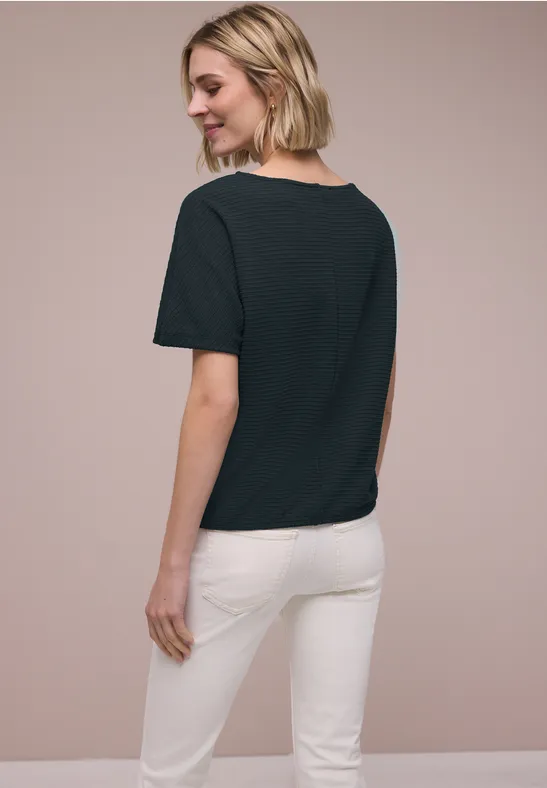 Vrouw van achteren in zwart T-shirt en witte broek voor een neutrale achtergrond.