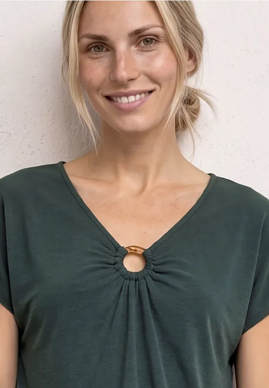 Vrouw met donkergroene korte mouw top met ringdetail bij de hals, staand voor een lichte muur.