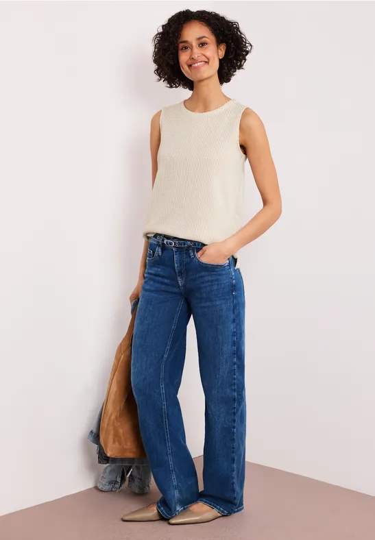 Vrouw in crèmetop en wijde spijkerbroek poseert nonchalant voor een muur.