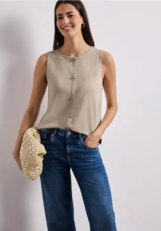 Eine Frau trägt eine ärmellose beige Bluse und blaue Jeans, hält eine geflochtene Tasche.