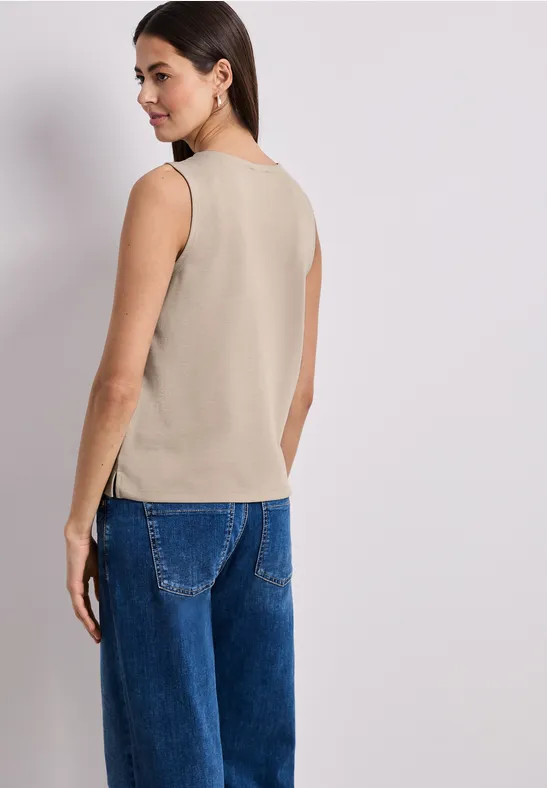 Frau steht mit dem Rücken zur Kamera, trägt ein beiges ärmelloses Top und weite blaue Jeans.