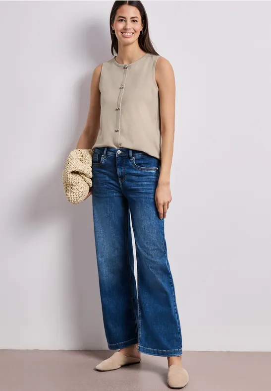 Frau in beige Top und weiten Jeans, mit Strohtasche am Arm und beigen Maultieren.